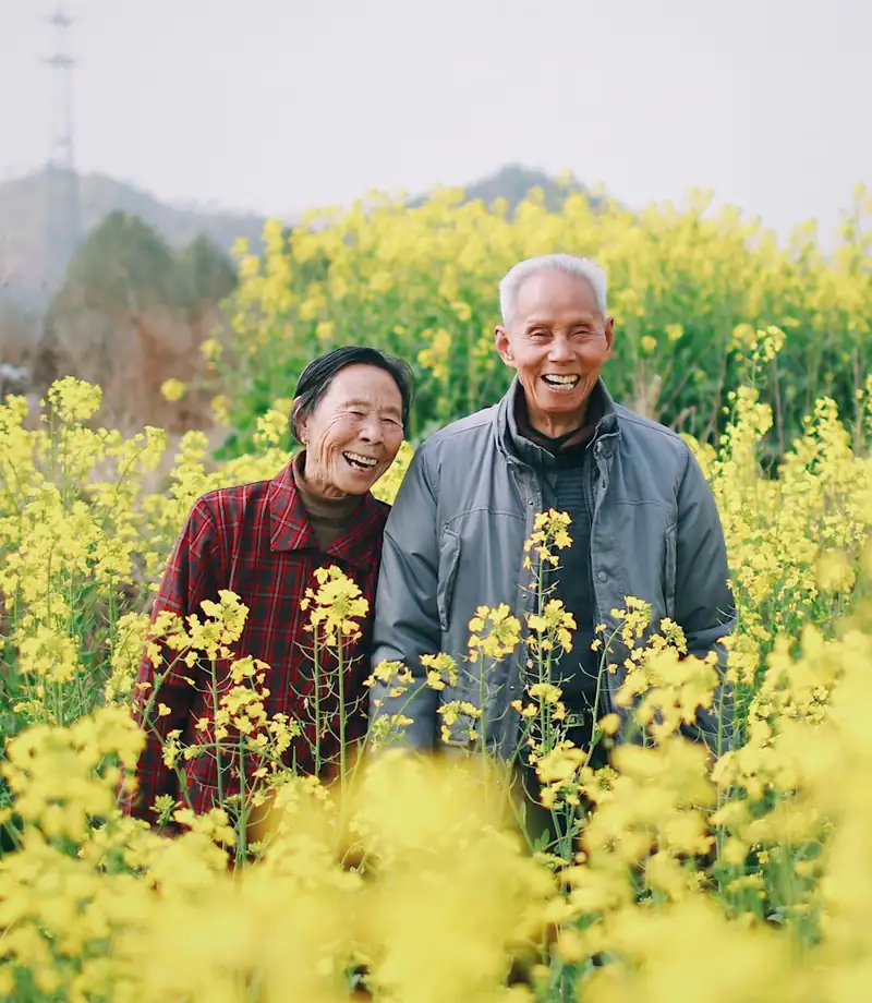 An elderly couple standing together outdoors surrounded by flowers